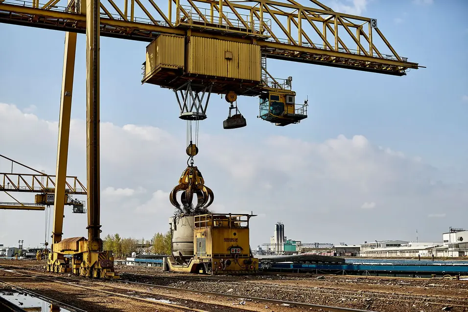 Gantry crane with a clamshell grab lifting scrap metal into a container in an industrial yard under a clear sky