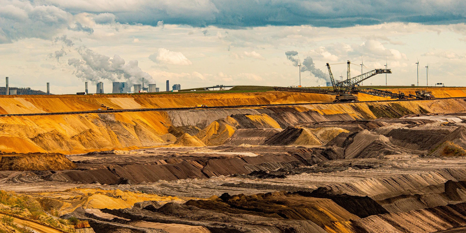 An open-pit mine with layered earth tones and large industrial machinery under a cloudy sky.