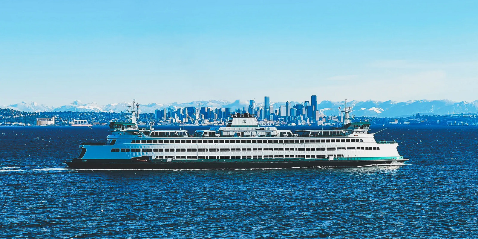 Ferry crossing a large body of water with a city skyline and mountains in the background.