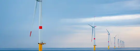 An offshore wind farm with a line of large wind turbines stands in the open water under a cloudy sky.