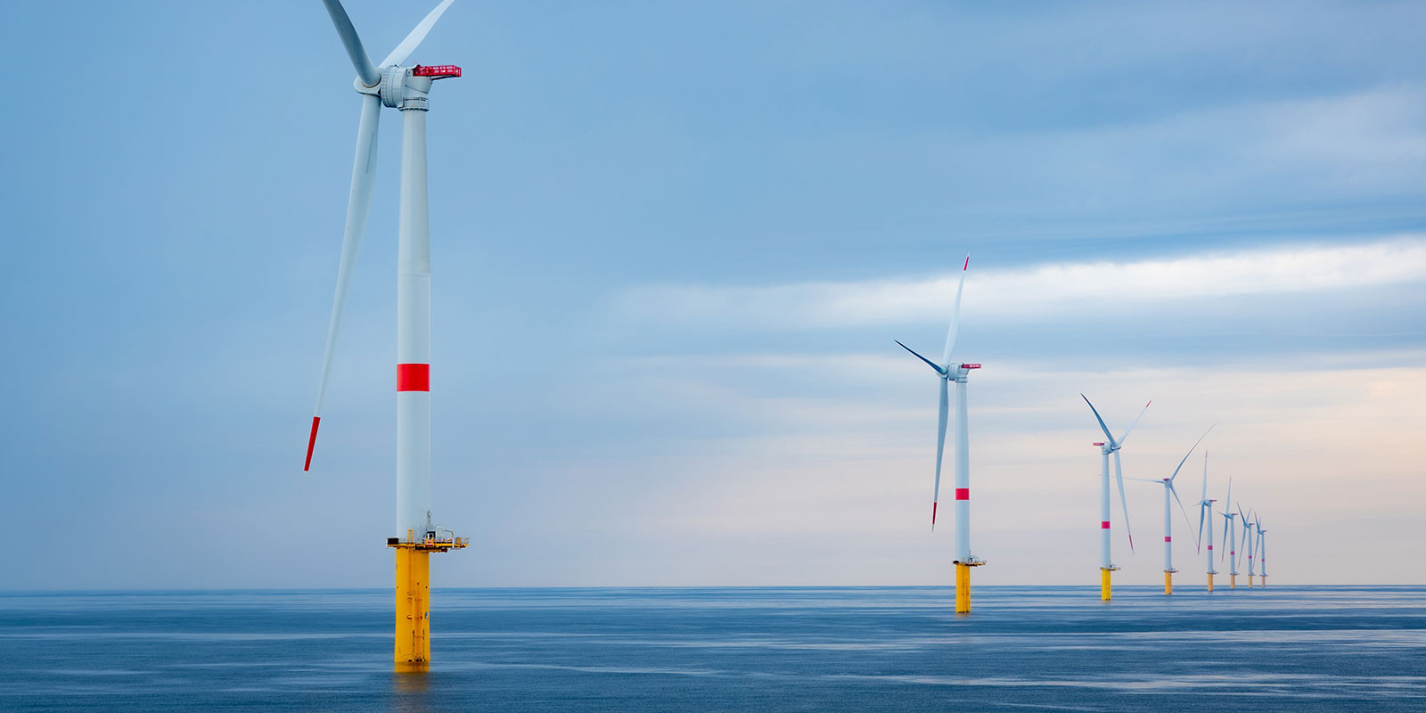 An offshore wind farm with a line of large wind turbines stands in the open water under a cloudy sky.