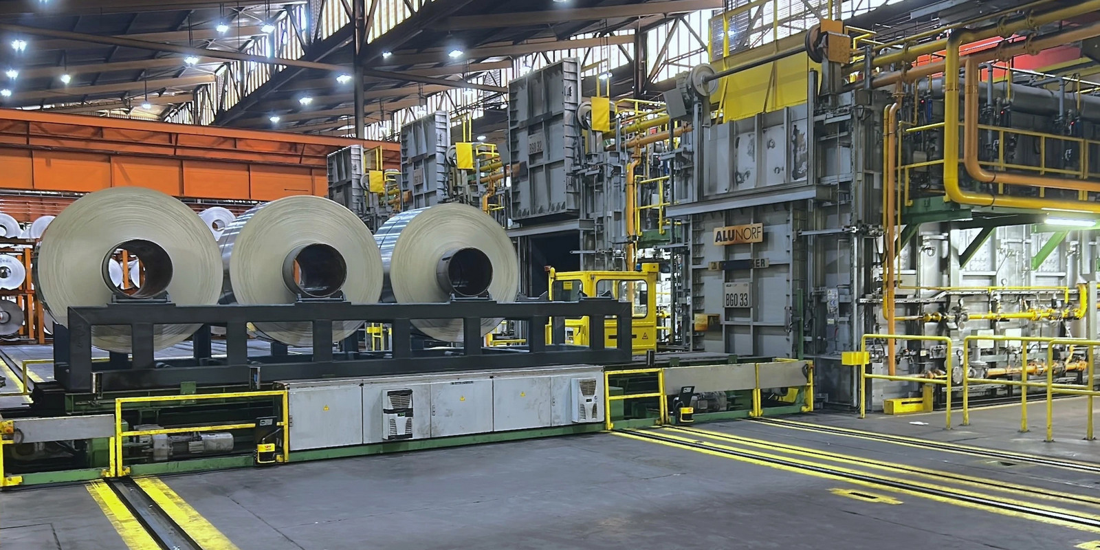 Large industrial facility interior with steel coils on a conveyor system and heavy machinery under bright factory lighting