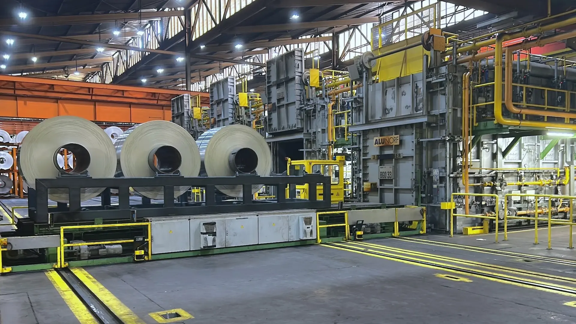 Large industrial facility interior with steel coils on a conveyor system and heavy machinery under bright factory lighting