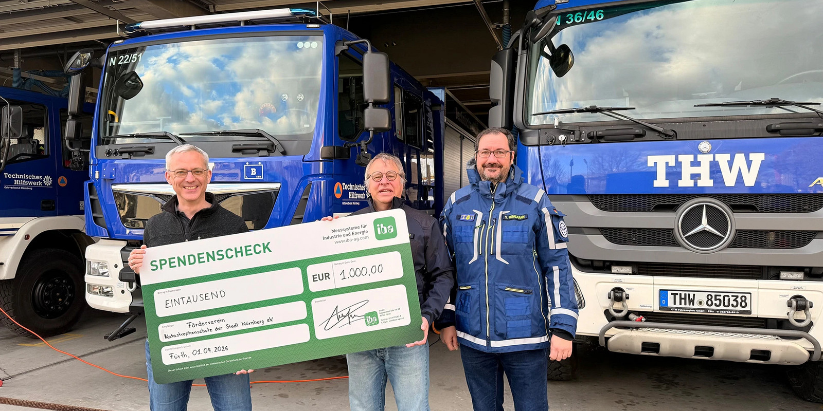 Three people stand in a vehicle hall in front of blue THW trucks, holding a large donation cheque for EUR 1,000.