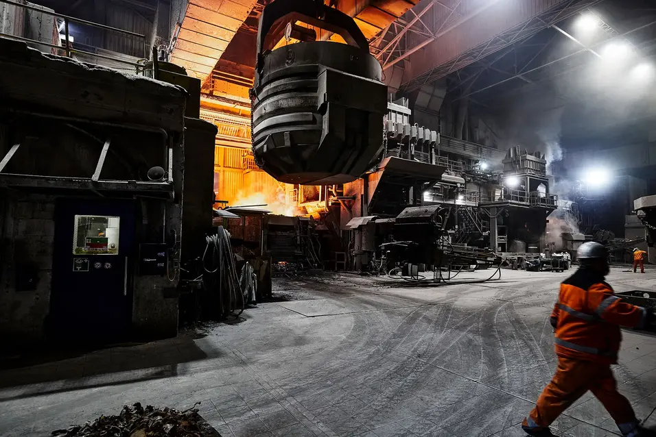 Large steel ladle suspended from an overhead crane inside an industrial hall with molten metal glow and a worker walking nearby