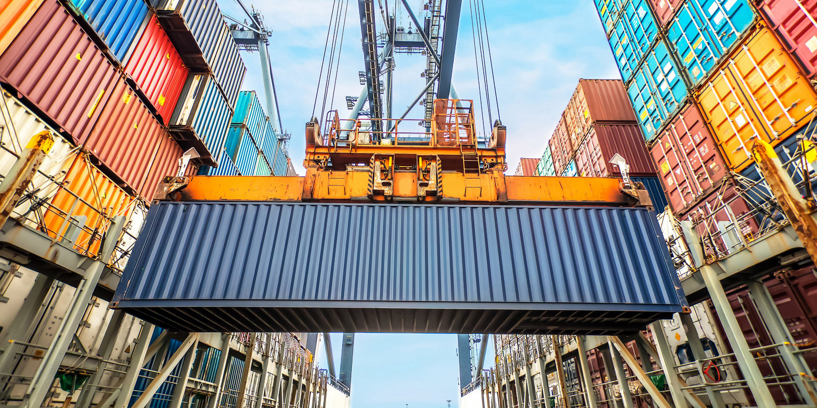 A large blue shipping container is lifted by a crane against a backdrop of colorful stacked containers.