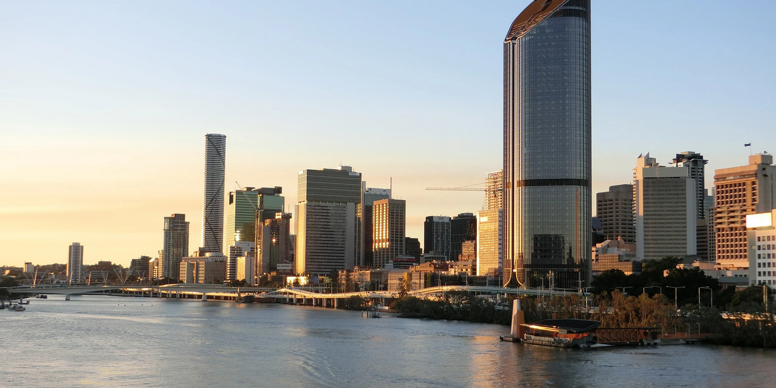 Brisbane skyline at sunset with modern high-rise buildings along the river and a curved glass tower reflecting warm light.