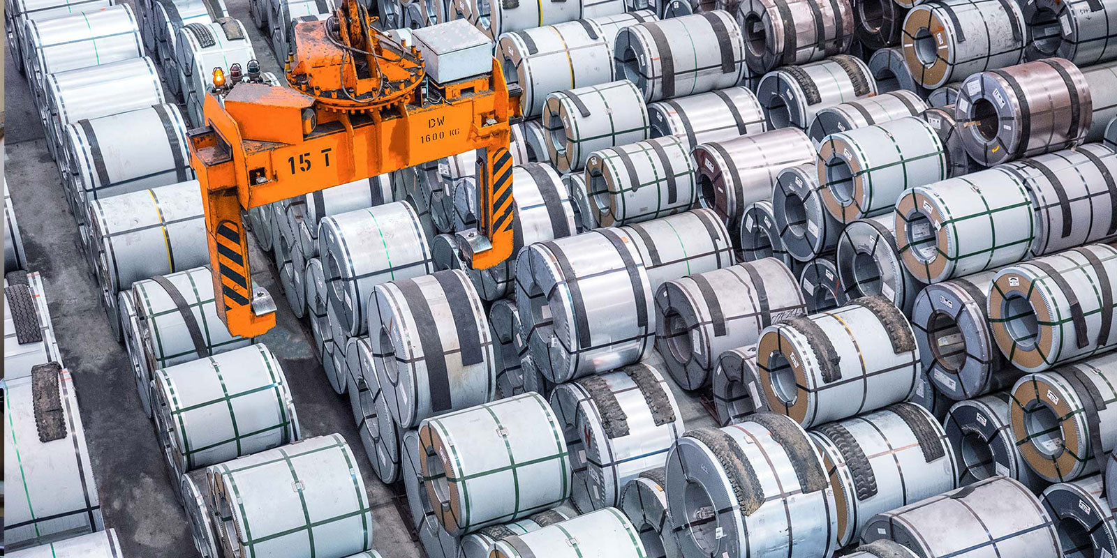 An orange 15-ton overhead crane is positioned above a massive collection of stacked steel coils.
