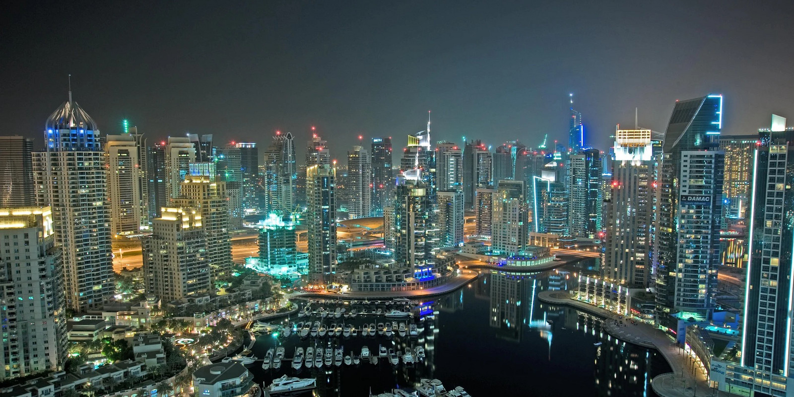 A night skyline with illuminated skyscrapers surrounding a marina filled with boats.